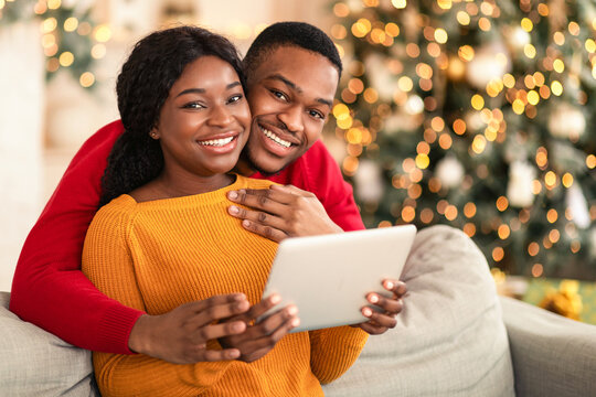 Online chat and congratulations to friends on new year during covid-19 lockdown. Young happy african american male and female use tablet in living room interior with Xmas tree with glowing garlands - Powered by Adobe