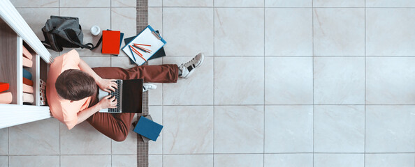 Student's Lifestyle Background. Guy Using Laptop Sitting Near Bookshelf On Floor In University Library. Top View, Panorama, Copy Space © Prostock-studio