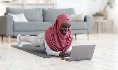 Home Sports. Active fit black muslim lady in hijab doing yoga plank exercise in front of laptop,...