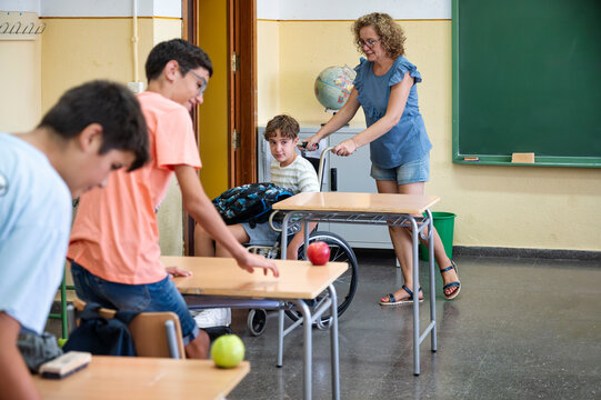 Teacher assisting student in wheelchair while classmates interact inside elementary classroom with desks and apples
