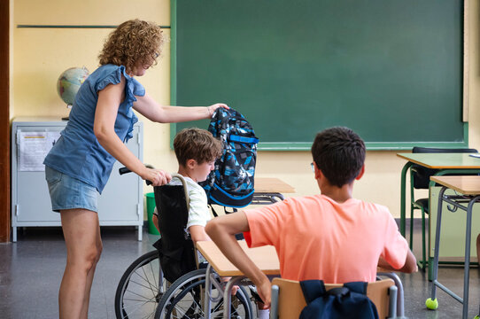 Teacher assisting elementary school student in wheelchair with backpack inside classroom