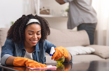 African Couple Making Spring-Cleaning At Home Together, Woman Wiping Table With Rag And Sprayer While Her Boyfriend Washing Shelves From Dust