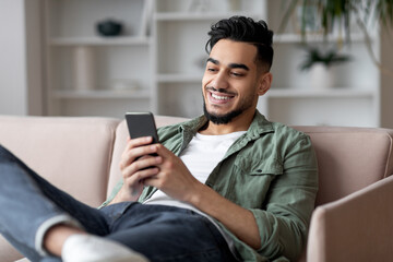 Portrait Of Handsome Young Arab Guy Relaxing With Smartphone At Home, Smiling Middle Eastern Male...