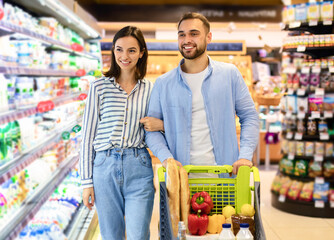 Happy Millennial Family Couple Buying Food In Supermarket, Choosing Products Walking With Cart...