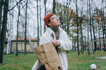 Stylish man wearing a white coat and orange beanie is holding a large paper bag and looking up while standing in a park during the autumn season