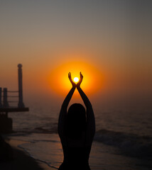 Silhouette of woman standing on beach holding sun with raised hands during sunrise. Ocean shore and pier at morning. Sunsrise and relaxation concept.