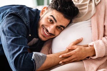 Future Father. Happy arab man listening his pregnant wife's belly, loving muslim guy embracing big...