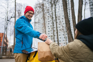 Smiling delivery man wearing a hat and jacket, carrying a thermal backpack, handing a fast food order to a customer in an urban park during the chilly winter season