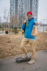 Smiling delivery man in a red hat and blue jacket, riding a longboard through a city park while offering a paper bag filled with a food order for a convenient meal