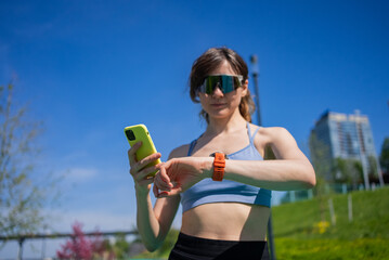 Young athletic woman checking progress on smartwatch and smartphone app after outdoor workout in city park, enjoying healthy lifestyle and achieving fitness goals under blue sky