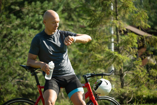 Sporty caucasian man and cyclist with smartwatch check apps, fitness progress at public park. Summer outdoor activity.