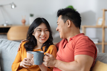 Closeup photo of happy loving asian couple sitting on couch in living room, drinking tea together,...