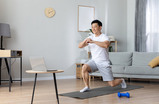 Motivated athletic handsome middle aged asian man exercising at home, training on yoga mat, using laptop, watching online fitness class, copy space. Workout plan for men concept
