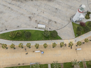 Aerial view of a white lighthouse with red accents standing tall against the gray concrete, bordered by lush green grass and palm trees, Batumi, Adjara, Georgia.