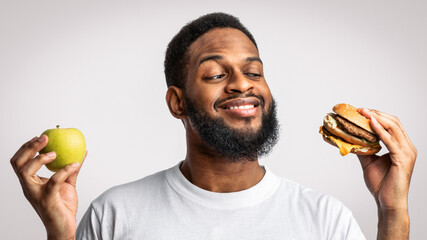 Black Guy Holding Burger And Apple Choosing Between Healthy Vs Unhealthy Food Standing In Studio On...