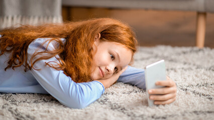 Spending Time At Home. Portrait of cal smiling red-haired genz tween girl using her mobile phone, watching video or movie online lying on the floor carpet in living room or bedroom