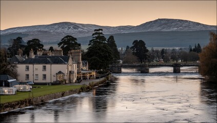 Obraz premium Scenic riverside town at dawn, with snow-capped mountains in the background. A bridge spans the river
