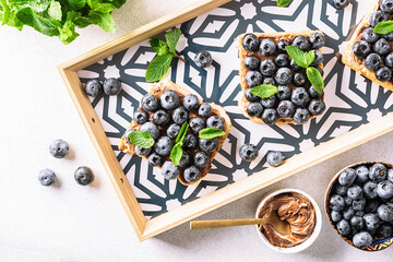 Sweet homemade toasts with chocolate paste, nuts, blueberry and aromatic mint leaves on the serving board on dark stone background. Tasty summer dessert.
