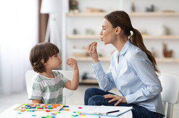 Young woman speech therapist studying together with small kid, learning practice pronunciation exercises with little boy child at office, free space