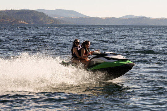 Two young women having fun on Couer D'Alene lake in Idaho riding the water on a fast jet ski watercraft outside on a long summer day. The sisters are laughing and enjoying the family time together. 