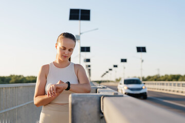 A woman in a white athletic top and smartwatch looks at her wrist, with a car approaching on a bridge lined with solar-powered streetlights.