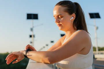 A peaceful woman with wireless earbuds looks down at her smartwatch, leaning on a bridge railing...