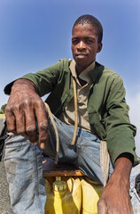 africa village, portrait of young african man , outdoors against the blue sky, sited on plastic drums with water