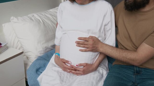 Medium midsection close-up of hands of caring unrecognizable man tenderly stroking belly of pregnant wife at maternity clinic, supporting during childbirth contractions and labor