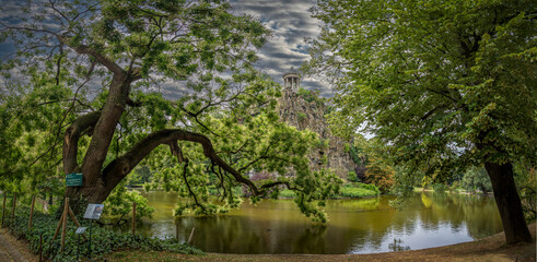 Paris, France - 07 27 2025: Park Buttes-Chaumont. Panoramic view of The Japanese Sophora tree, the belvedere island, Temple of the Sibyl and the lake behind