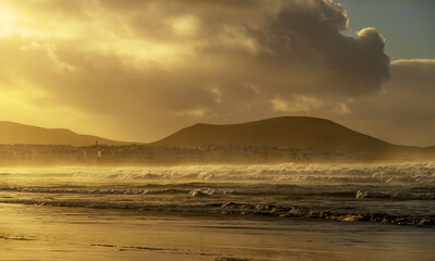Majestic clouds roll over Famara Beach on Lanzarote, creating a dramatic scene against the striking cliffs. Vast ocean waves touch the golden sand, enhancing the scenic beauty