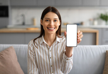 Joyful young woman in casual striped shirt presenting mobile phone with white blank screen for copy...