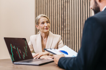 Businesswoman in a beige suit engaged in a meeting, analyzing financial data on a laptop, while a colleague presents a report with charts and graphs in a modern office setting