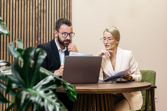 Business professionals engaged in strategic planning session, analyzing charts and financial data on laptop, surrounded by modern office decor and greenery, fostering collaboration and innovation