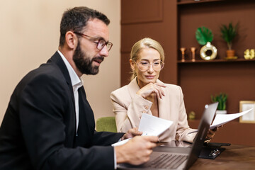 Business professionals engaged in a collaborative meeting, analyzing documents and discussing strategies while seated at a modern workspace with a laptop and decorative elements