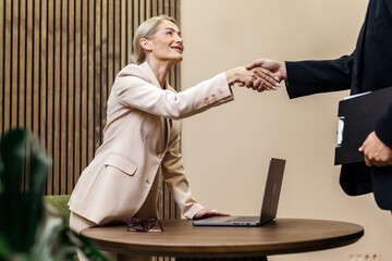 Businesswoman in a light suit shakes hands with a colleague at a modern office table, showcasing a...