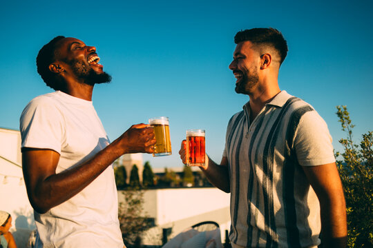 Two friends laughing and toasting with beer on rooftop at sunset