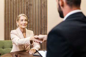 Businesswoman in a light suit shakes hands with a man in a dark suit at a modern office table,...