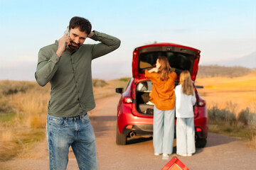 Man calling on cellphone, standing in front of broken car on the way to vacation, woman and girl...