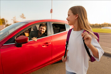 Happy schoolgirl with backpack going to school and smiling to her parents sitting in car, father...