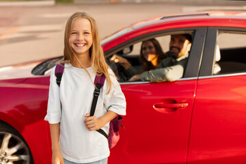 Ready for school. Portrait of happy European girl carrying backpack while standing next to car with parents sitting inside, arriving at school