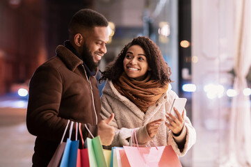 Choosing Present. Smiling african american woman showing her mobile phone to man, pointing at device, guy holding shopping bags, standing outdoors in the evening. Offer, Retail And Purchase