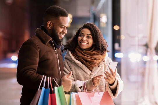 Fototapeta Choosing Present. Smiling african american woman showing her mobile phone to man, pointing at device, guy holding shopping bags, standing outdoors in the evening. Offer, Retail And Purchase