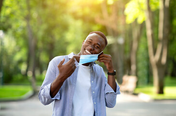 End of coronavirus pandemic. Happy African American guy taking off his protective mask at summer...