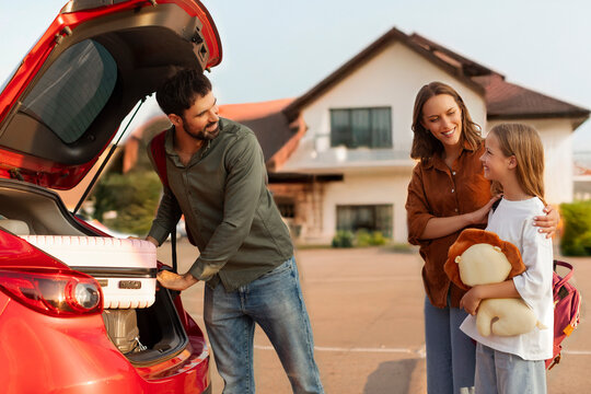 European family of three packing luggage into car truck in front of house, parents and daughter preparing to go on vacation