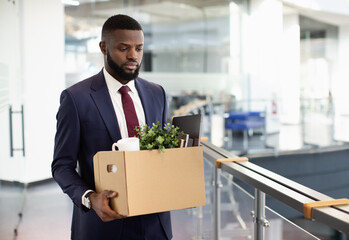 Sad young black man got fired, walking by office building and holding box with his belongings, copy space. Terminated african american guy in suit lost his job, leaving workplace, copy space