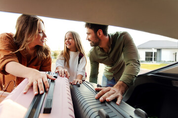 Family of three leaving for vacation, loading luggage into car trunk and smiling at each other, shot from inside vehicle