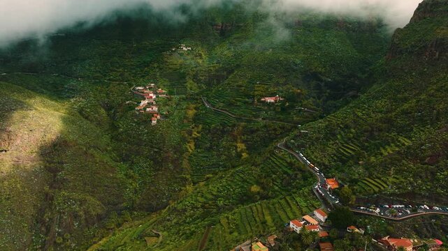 Aerial drone view over Masca village, terraced green mountains shrouded in clouds, scattered houses along winding road in the Canary Islands