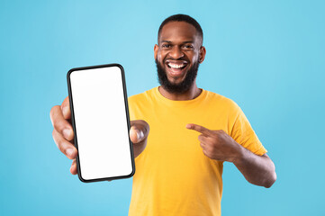 Handsome happy black guy pointing at smartphone with blank screen on blue studio background, mockup...