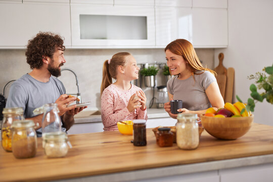 A family is sharing breakfast in a bright kitchen. The father holds a plate, while the daughter and mother engage in friendly conversation over fruit and homemade treats, enjoying their time together.
