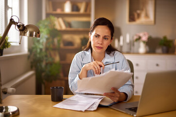 A woman sits at a desk in her home office, examining financial documents with a look of concern. A...